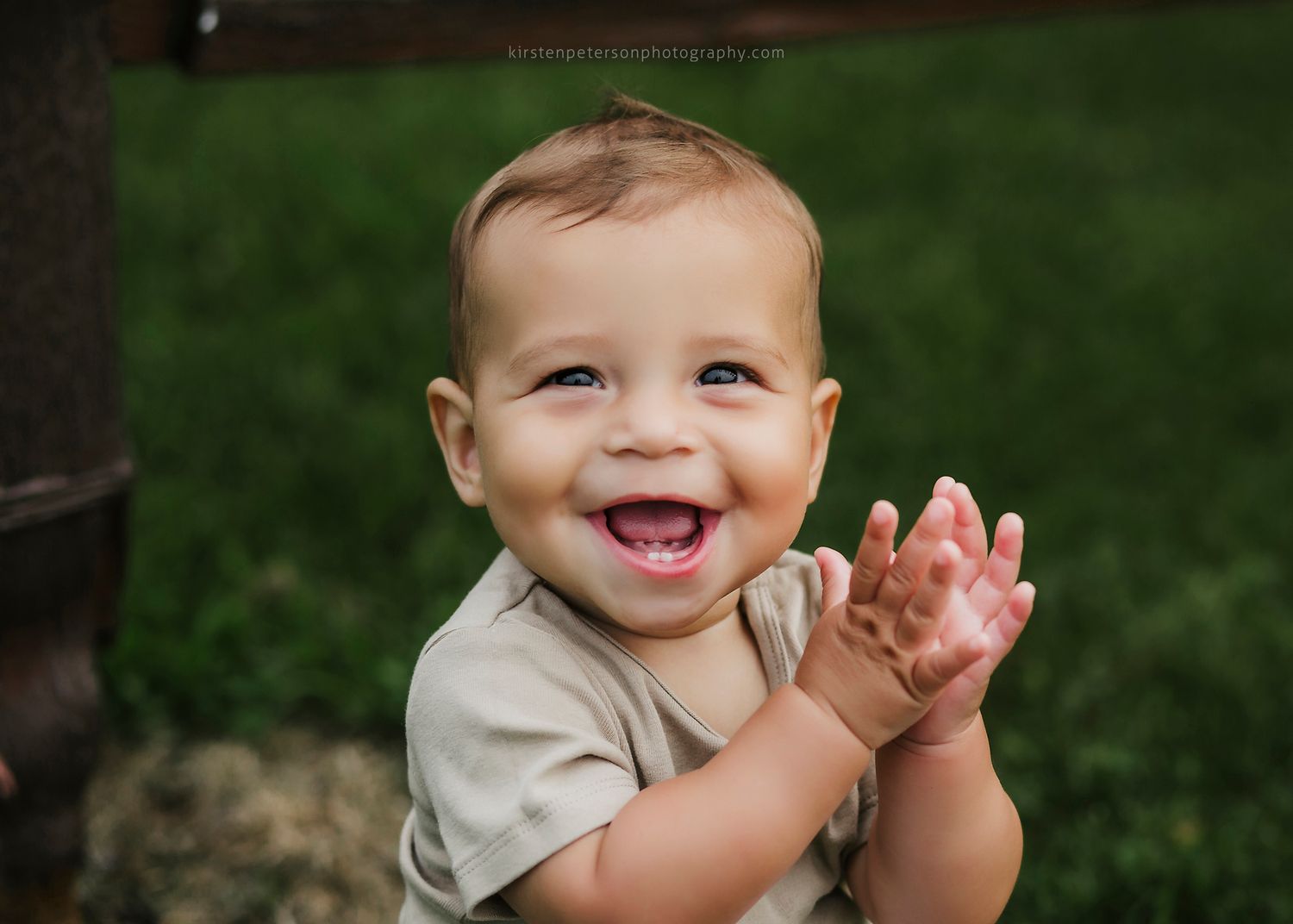 A happy baby wearing a light colored shirt sits outdoors in green grass while clapping and laughing.