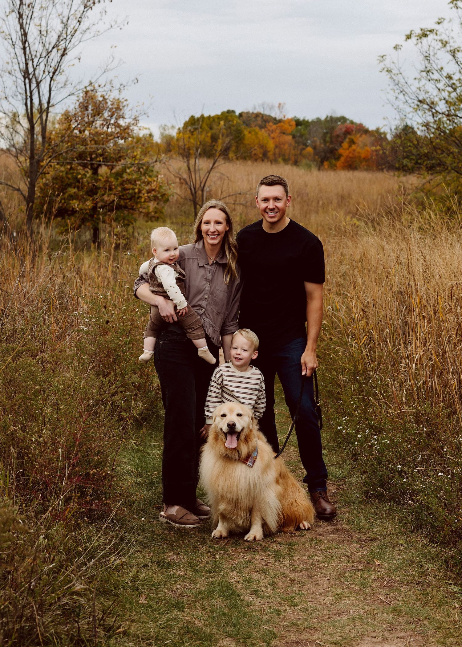 A family and their fluffy white dog pose together in a rustic outdoor setting with tall grass and autumn foliage.