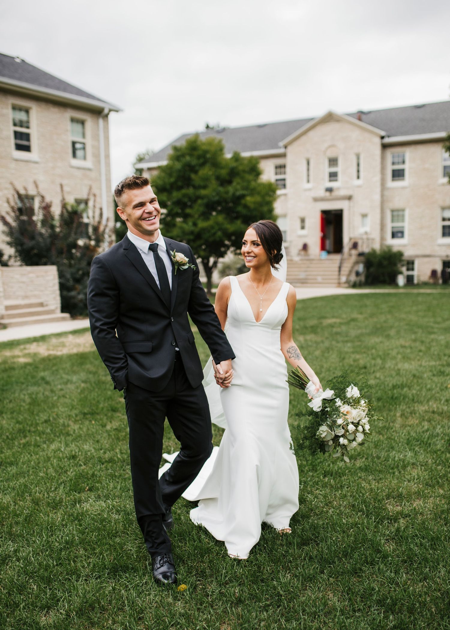 A couple shares joyful moments on a grassy lawn with residential buildings in the background during a wedding photoshoot.