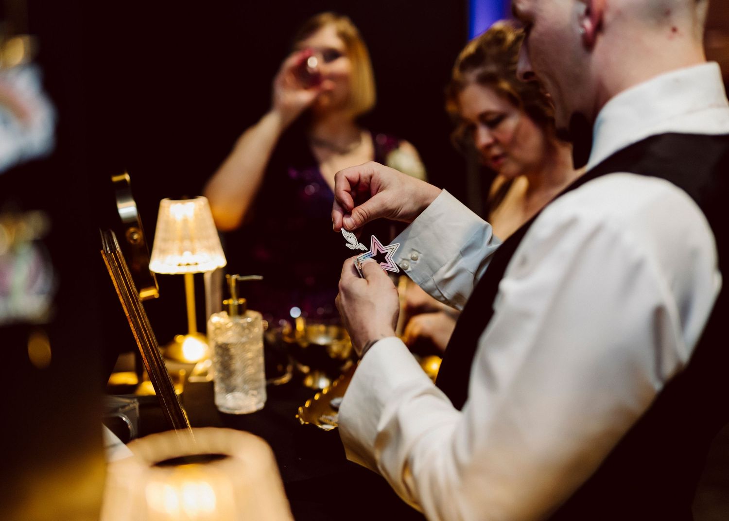 Bartender adjusting cufflinks at dimly lit bar with guests in background at elegant evening event.