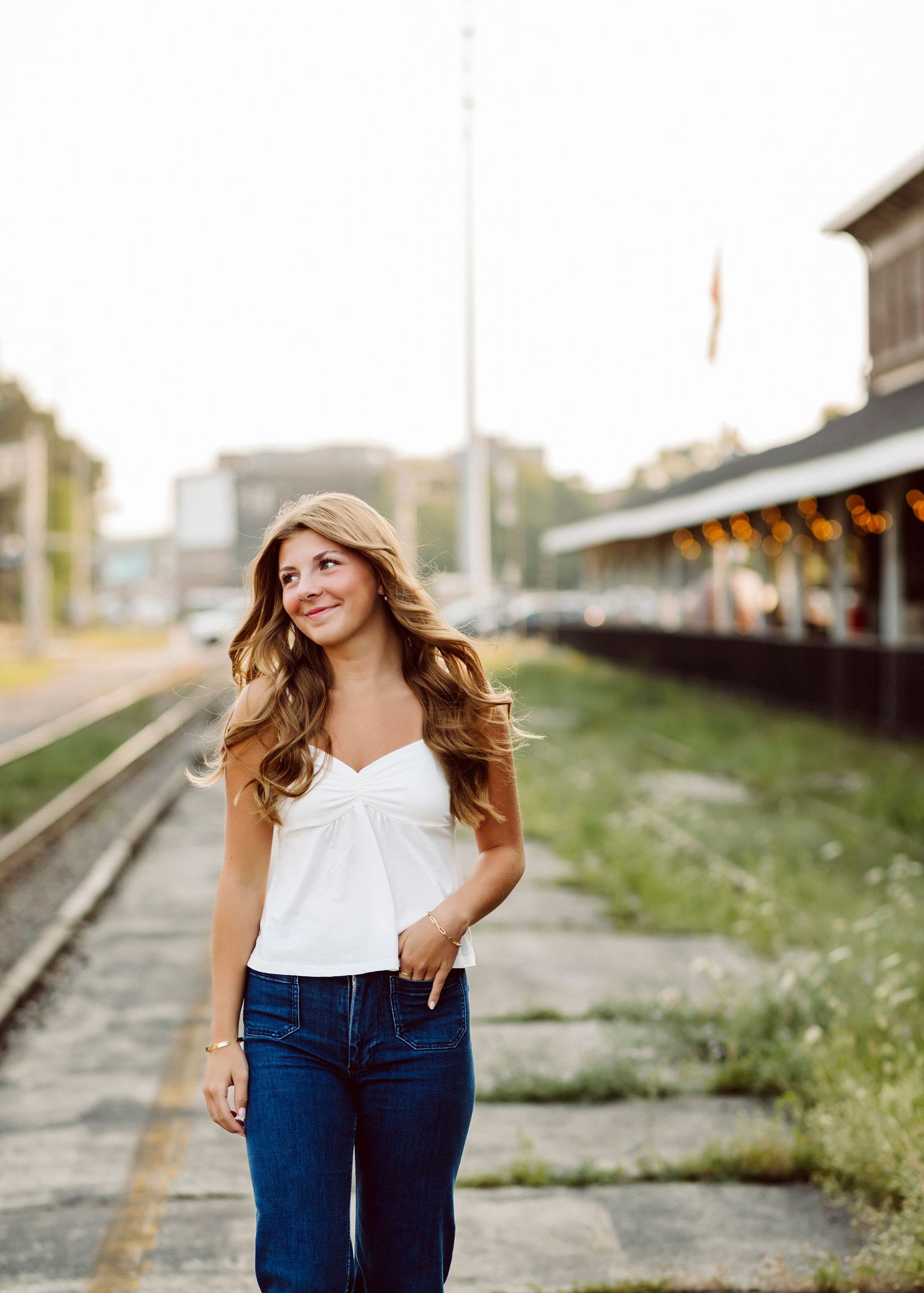 Woman in white sleeveless top and blue jeans poses near train tracks during golden hour.