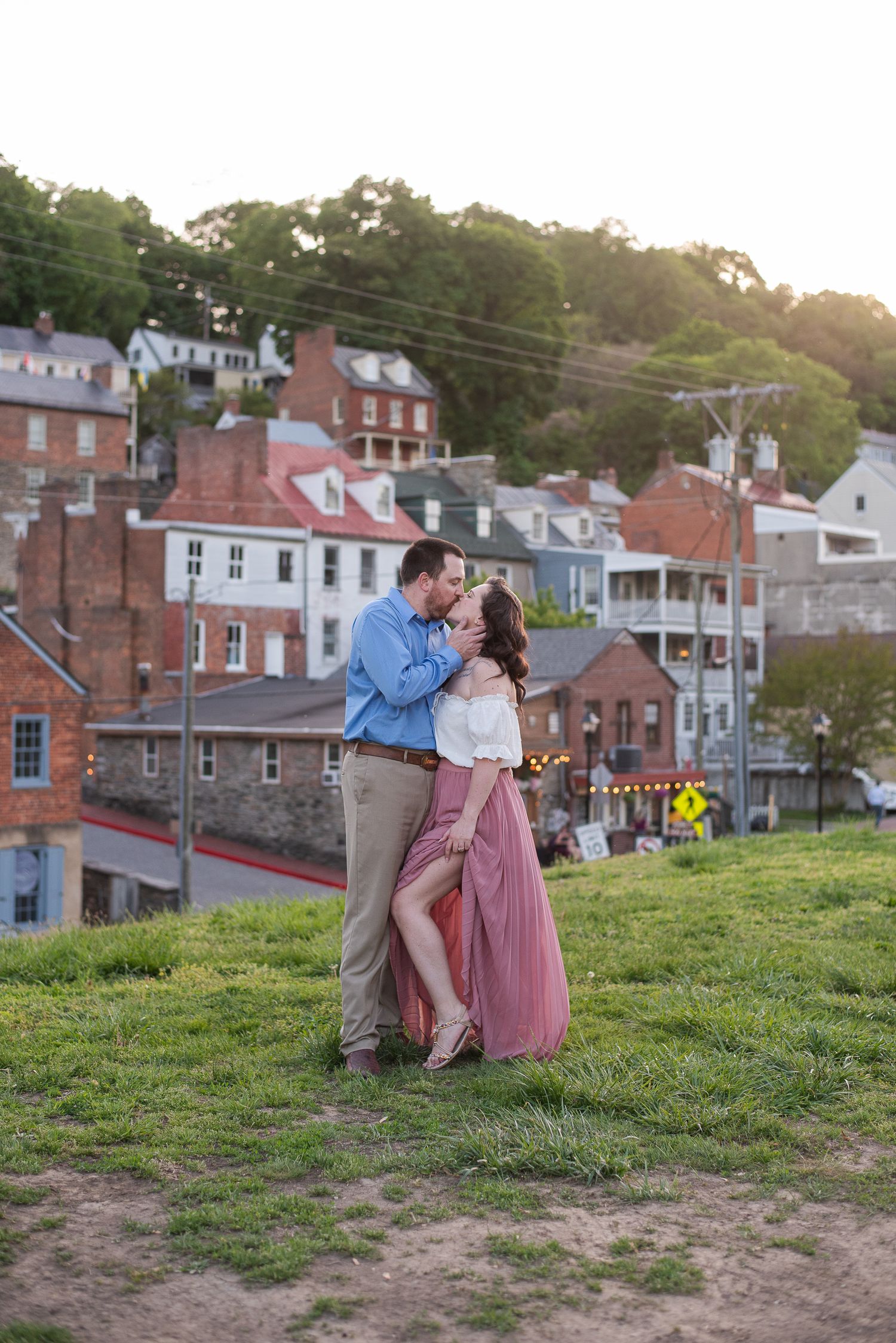 A couple embraces romantically on a grassy hillside overlooking historic brick buildings and houses at sunset.