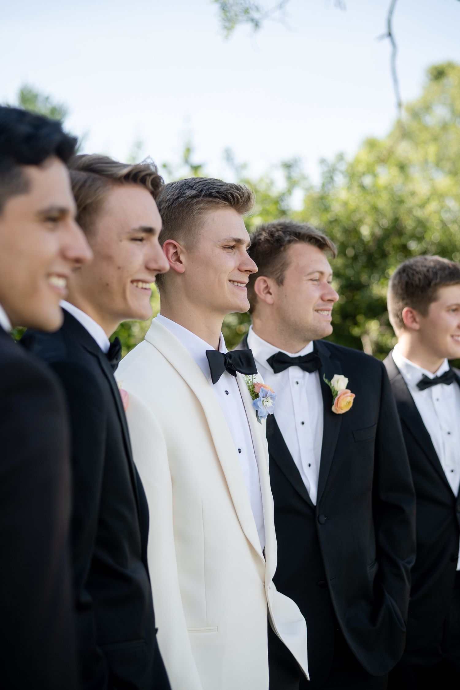 Groomsmen in formal black tuxedos and one in white stand together smiling outdoors at a wedding.