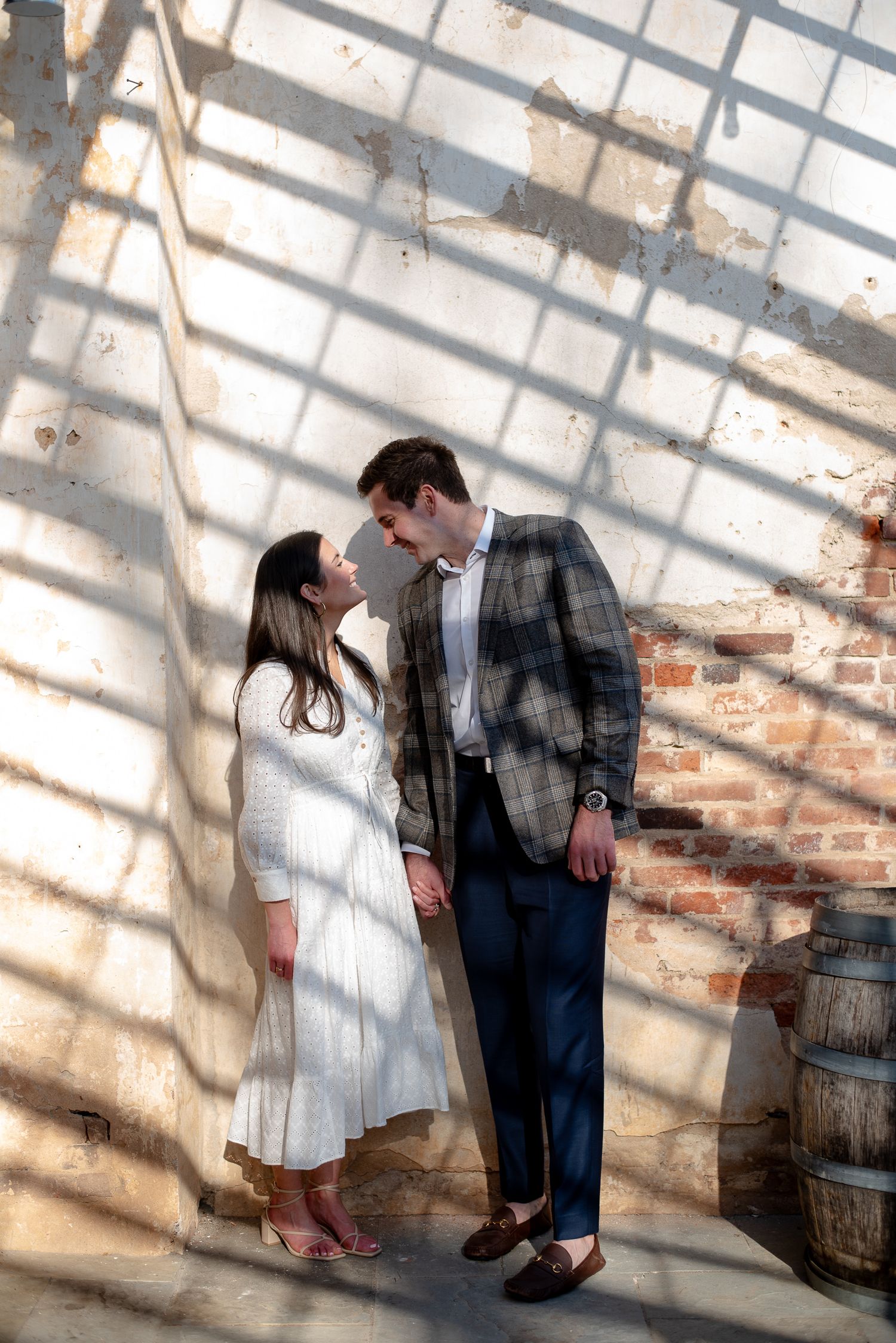 A couple shares an intimate moment against a sunlit brick wall with dramatic window shadow patterns.