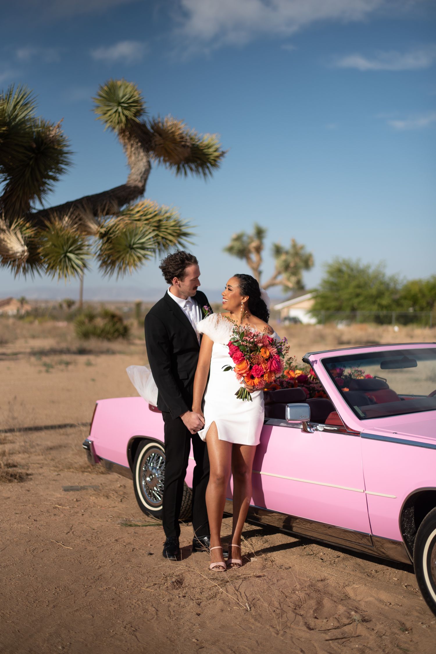 A couple celebrates near a pink vintage convertible car in a desert landscape with palm trees against a blue sky.