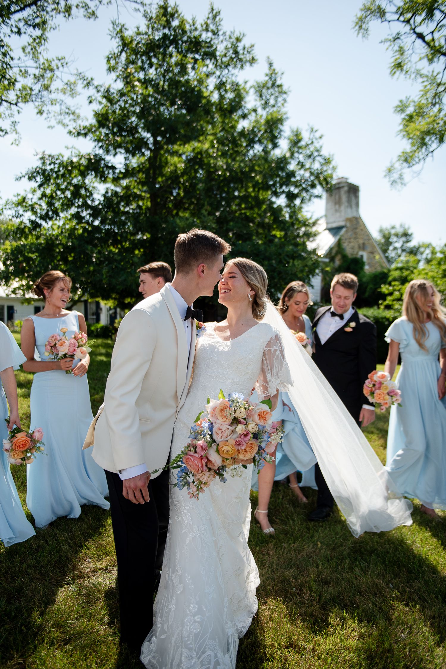 Wedding party in light blue and white attire pose playfully in a sunny garden setting.