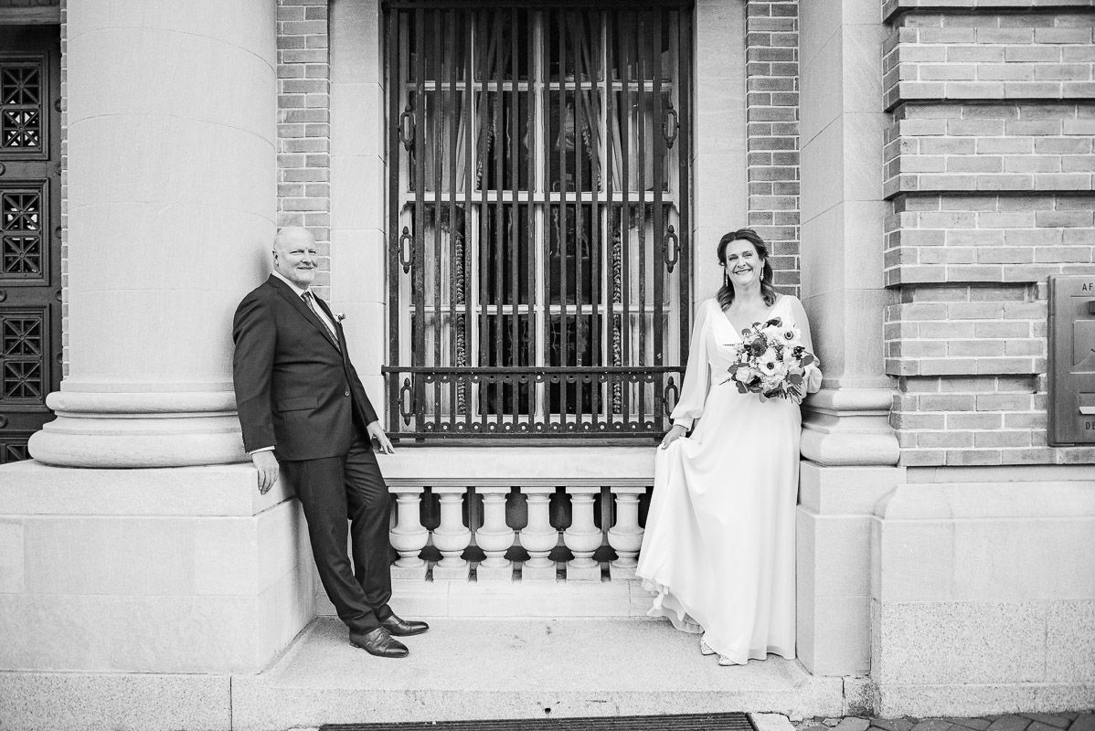 A couple poses against a classic stone building facade with ornate iron window details.