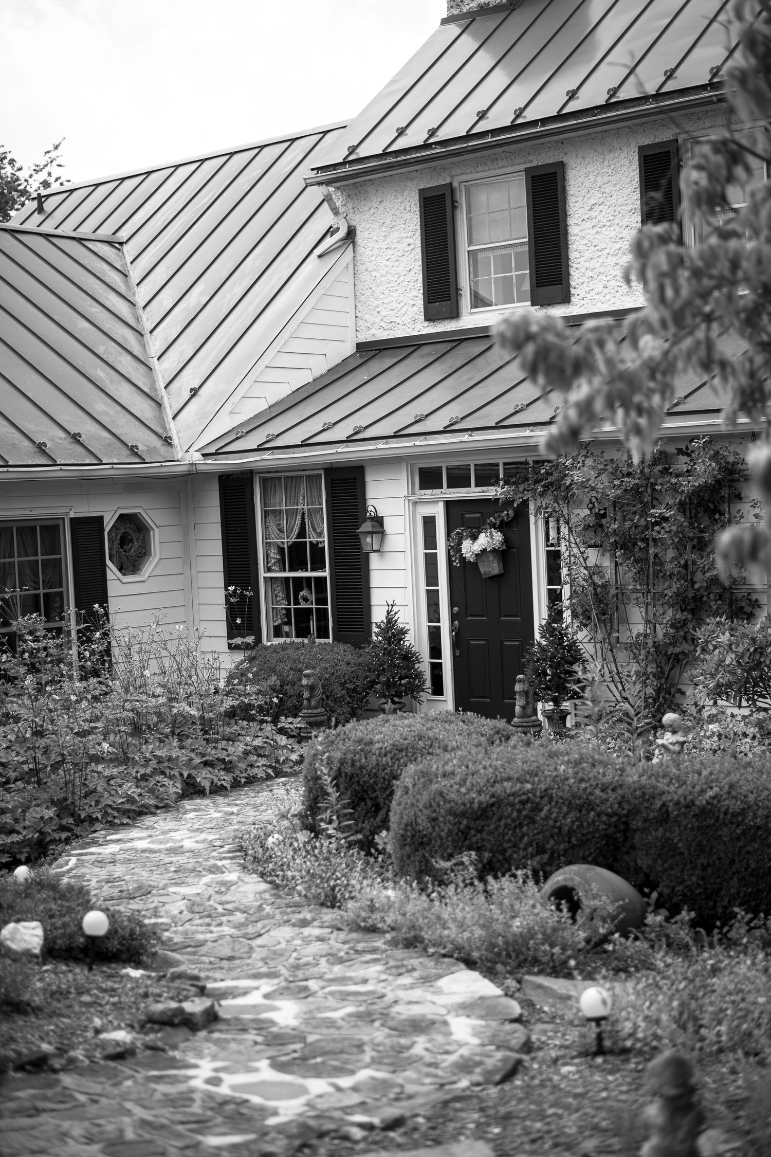 A charming cottage house with stone pathway through manicured garden and metal roof in black and white photography.