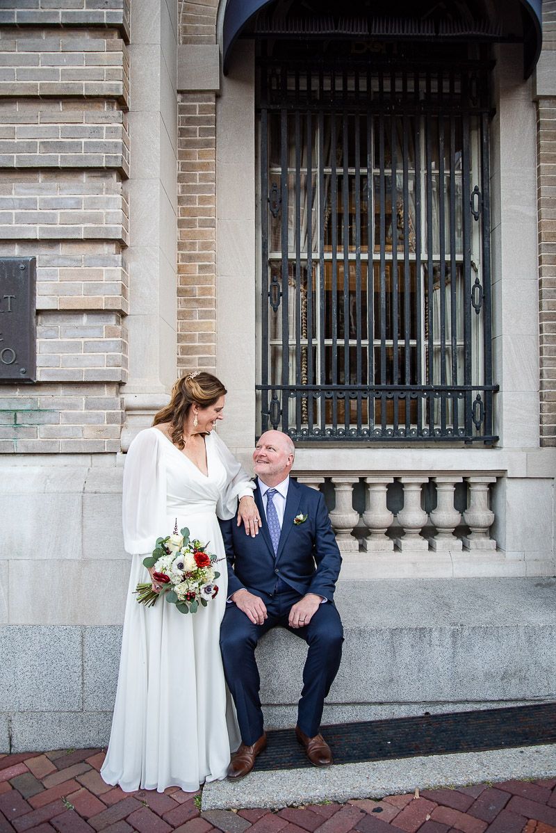 A couple in wedding attire shares intimate moments against a brick building with ornate windows.