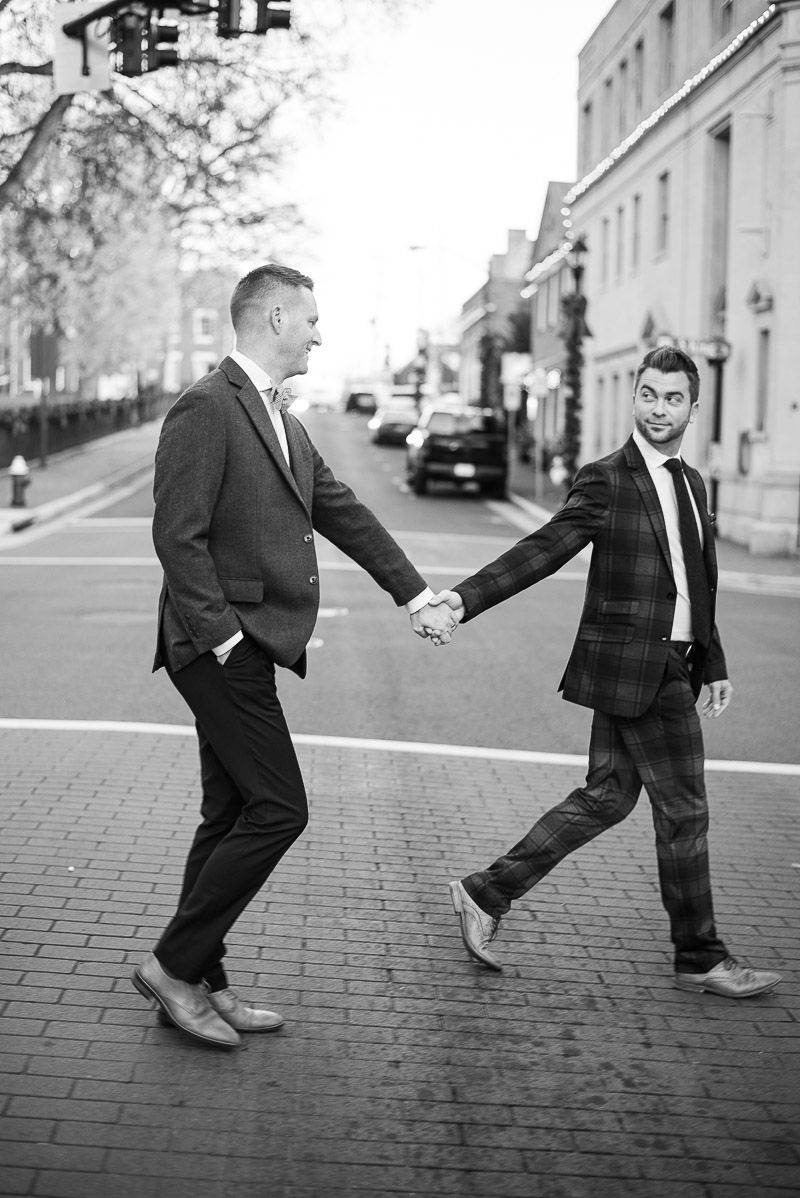 Black and white sequence of two men in suits playfully dancing and interacting on a brick sidewalk in an urban setting.