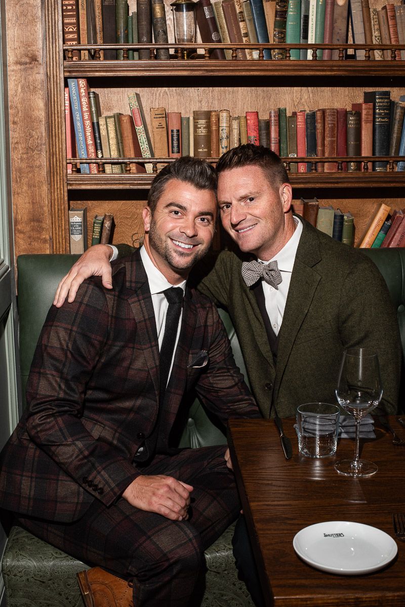 Two people in suits pose together at a bar with bookshelves in the background.