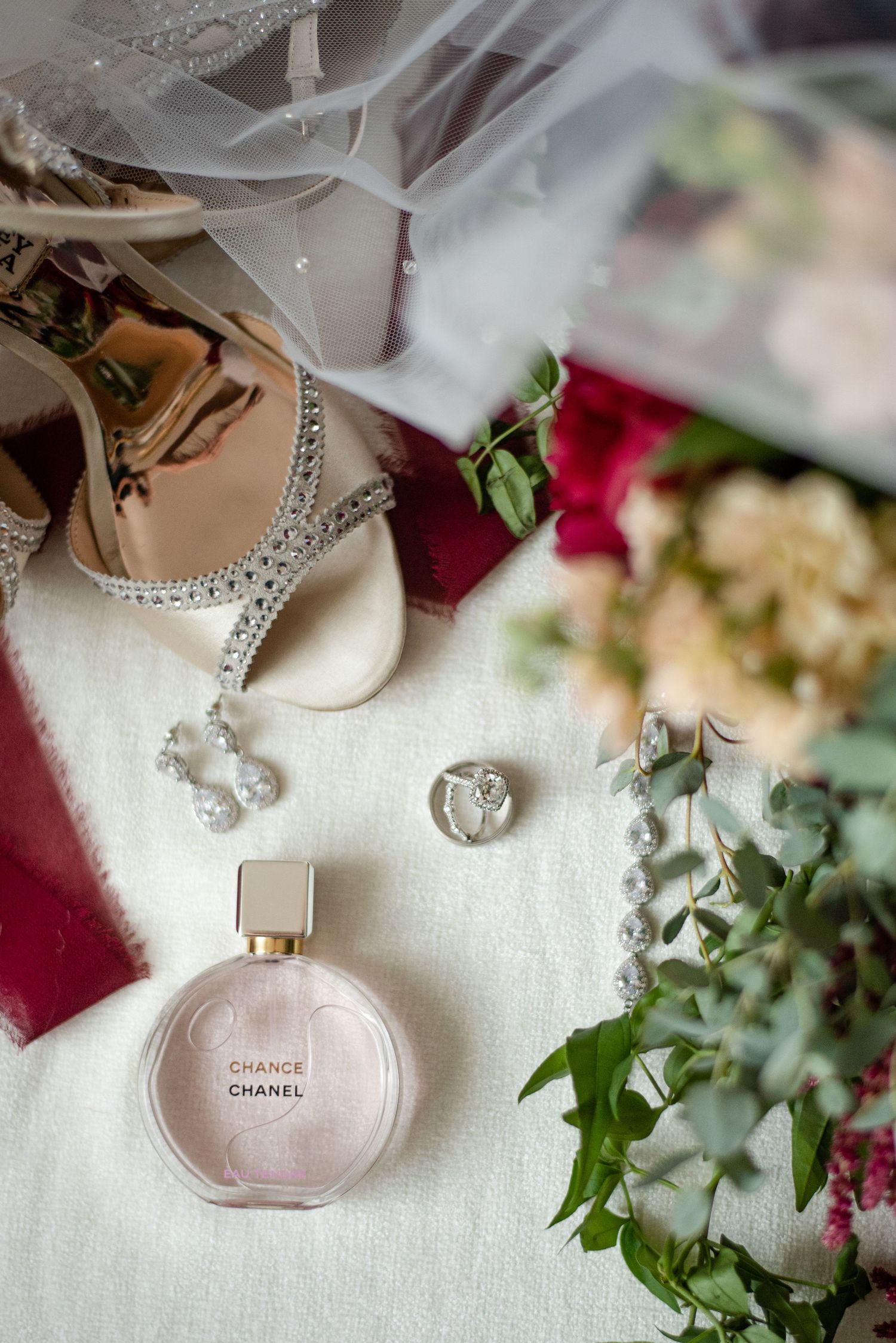Wedding accessories flatlay with Chanel perfume, jewelry, and floral details on white fabric.