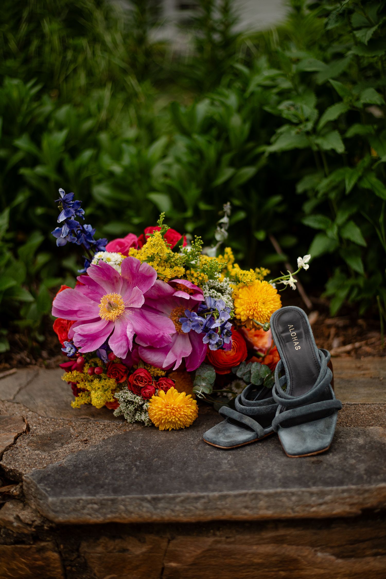 A vibrant wildflower bouquet with pink dahlias and yellow blooms sits beside gray leather sandals on a rustic stone surface.