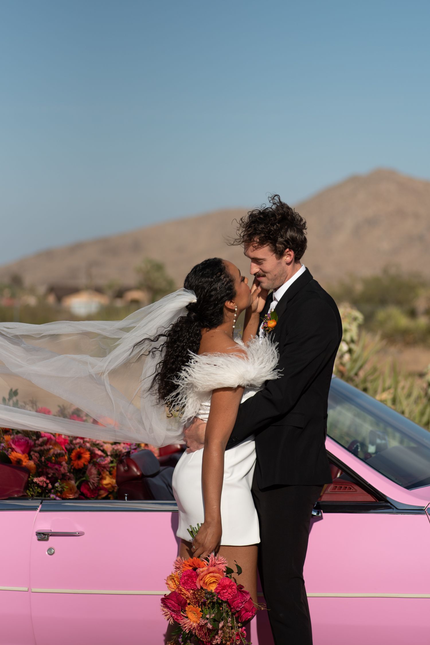 A romantic newlywed couple embrace beside a pink vintage car decorated with vibrant red and orange flowers in a desert setting.