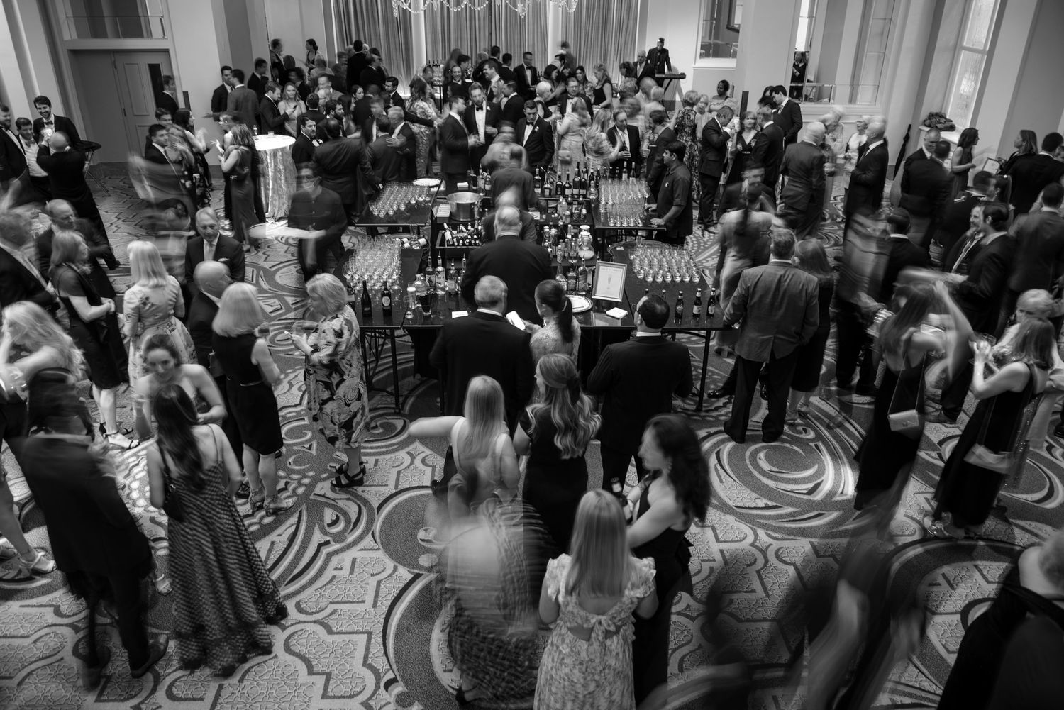 Black and white overhead view of cocktail hour with guests around the bar on patterned ballroom floor.