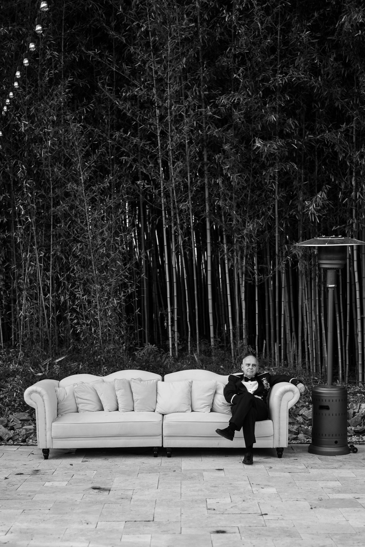 Stylish white couch placed outdoors against tall bamboo forest backdrop with someone lounging in formal attire.