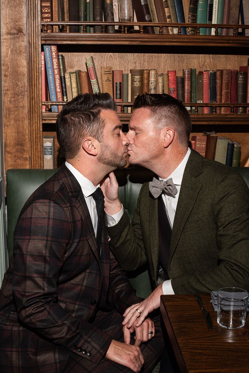 Two people share an intimate moment in a cozy library setting with wooden bookshelves in the background.