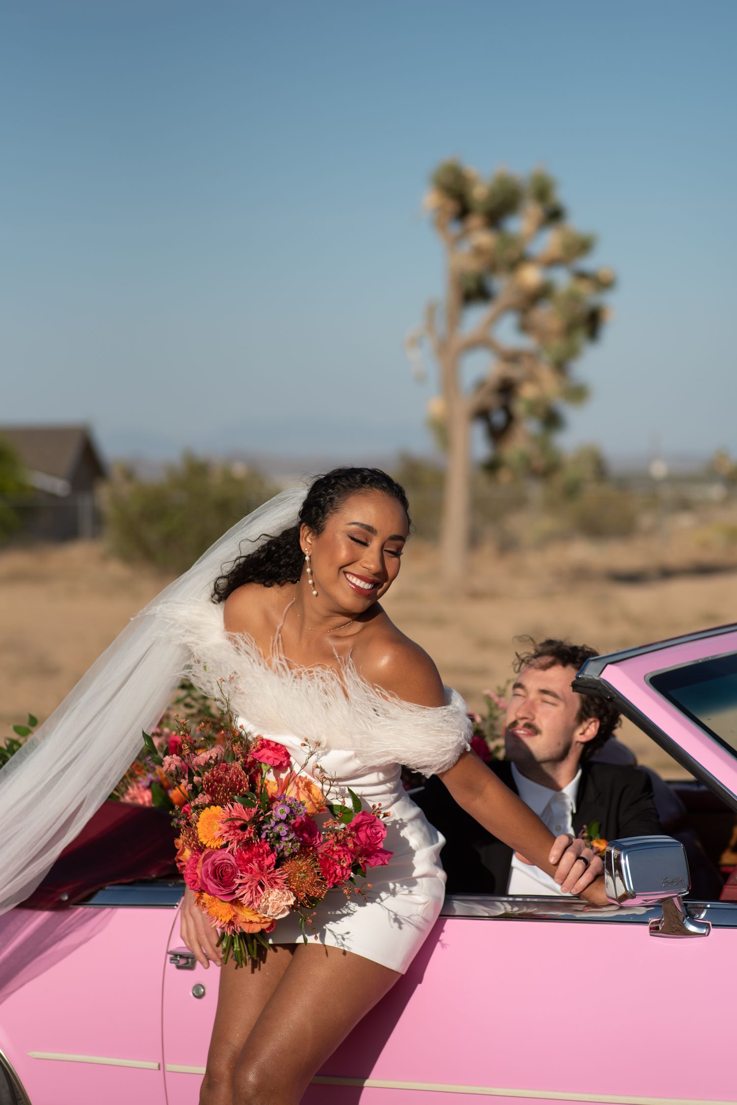 A wedding scene in the desert with a pink vintage car, vibrant bouquet and Joshua tree in the background.