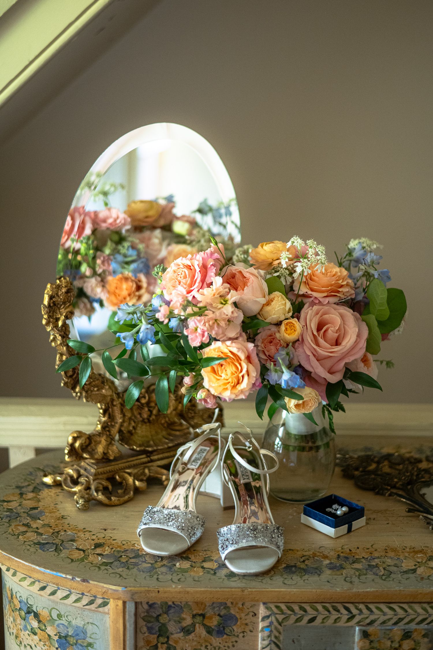 Elegant bridal shoes and colorful floral arrangement with pink roses displayed on ornate vintage table with decorative mirror.