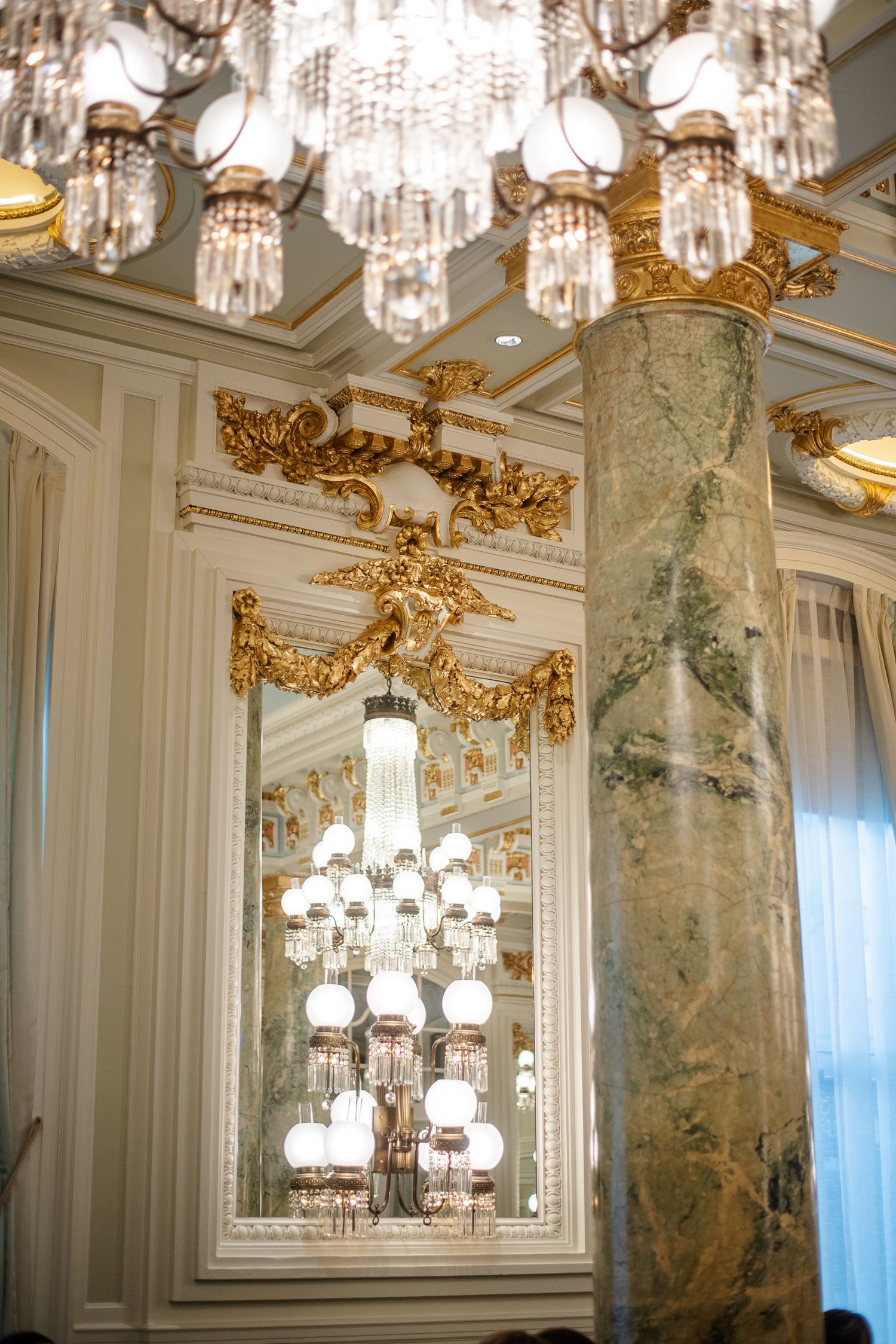 Ornate crystal chandeliers and gilded decorative elements adorn a luxurious room with marble columns and mirrored walls.