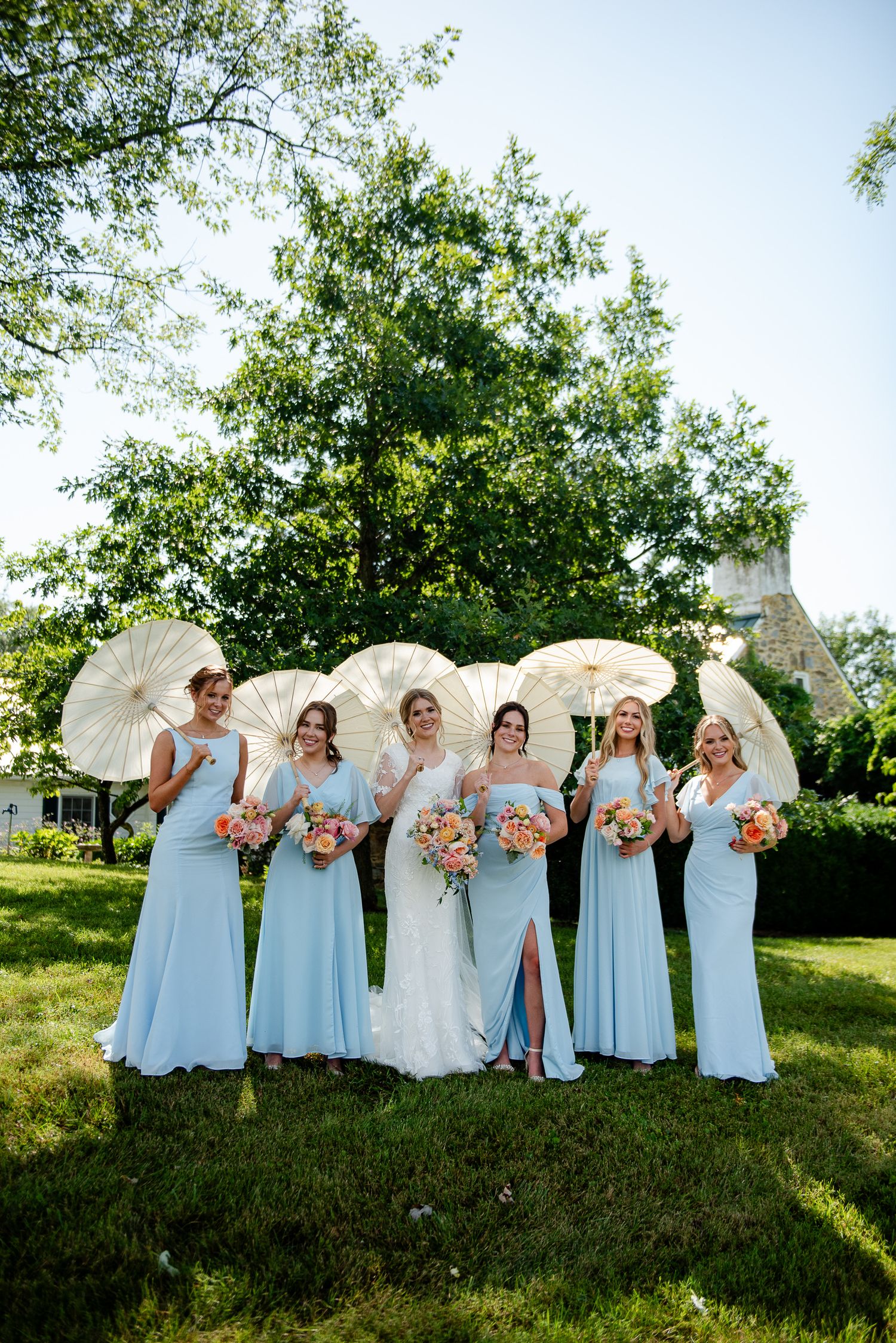 Wedding party with bridesmaids in light blue dresses holding white parasols and bouquets pose outdoors on sunny summer day.