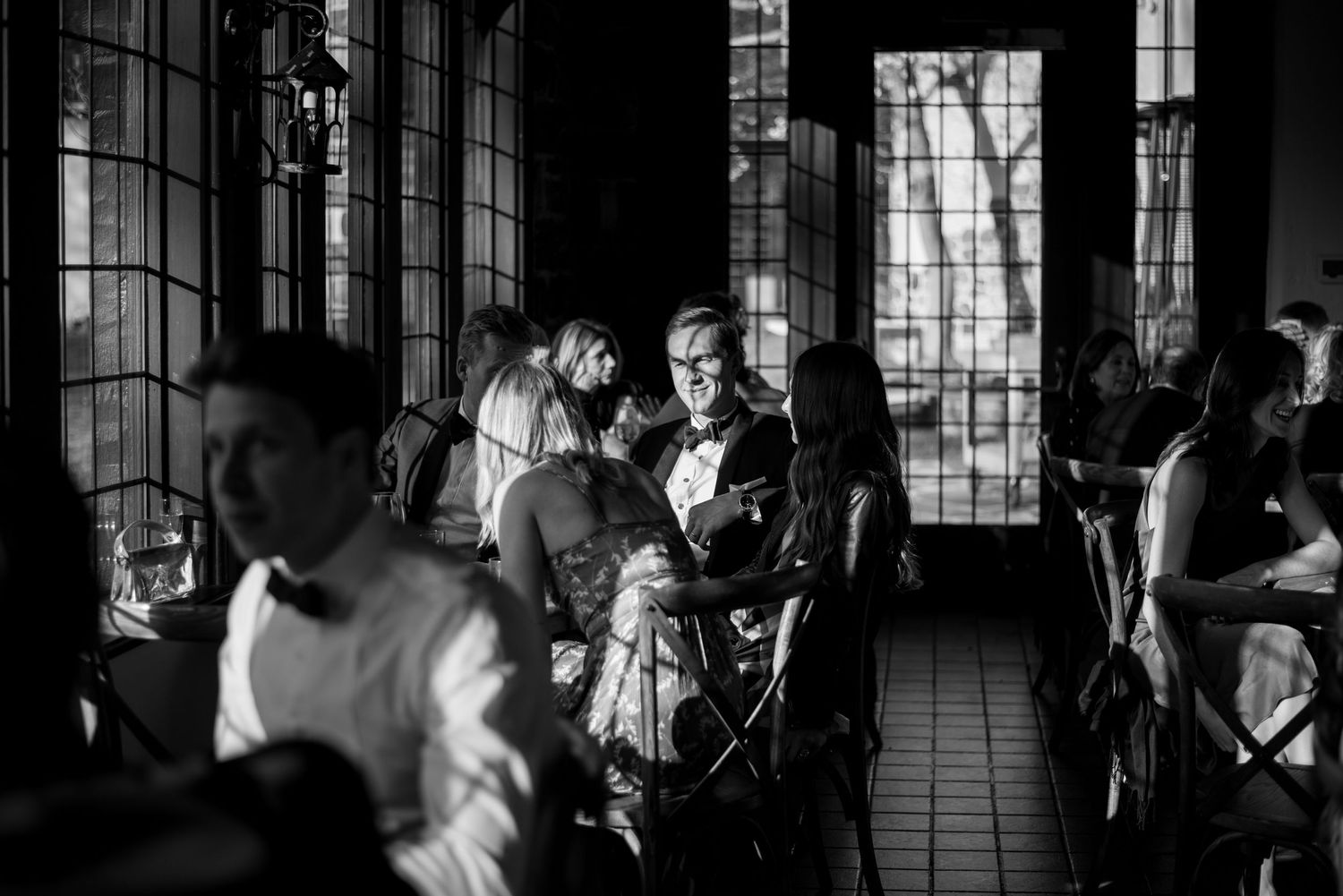 Black and white photo of guests seated during cocktail hour with tall windows.