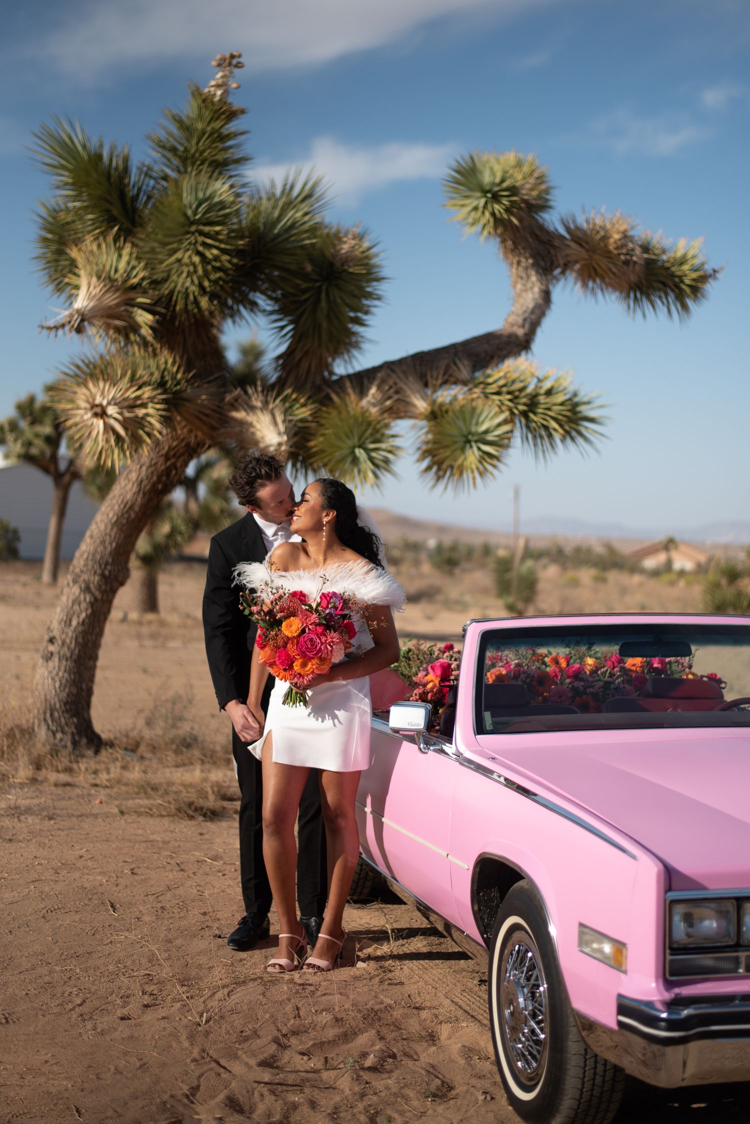A vintage pink Cadillac parked in a desert landscape with Joshua trees under a blue sky.