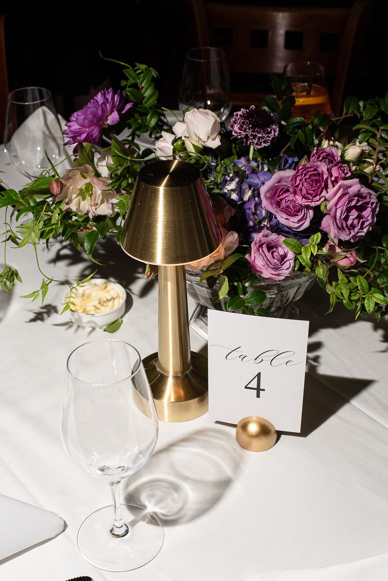 Elegant table setting with brass lamp, purple floral arrangements, and table number card on crisp white tablecloth.