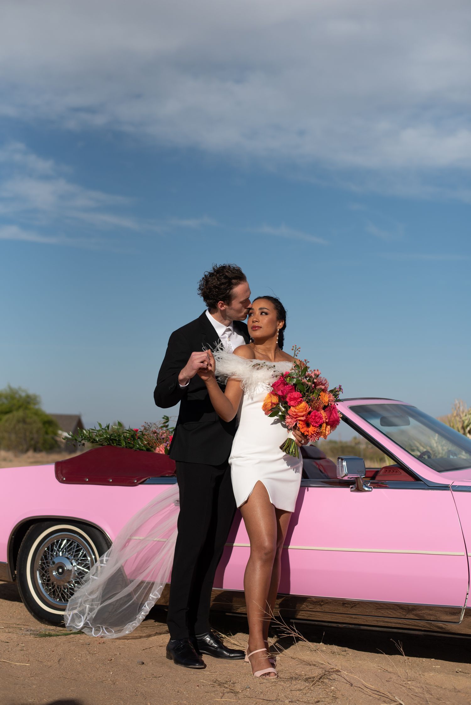 A couple poses together against a pink vintage car in the desert with a colorful bridal bouquet.