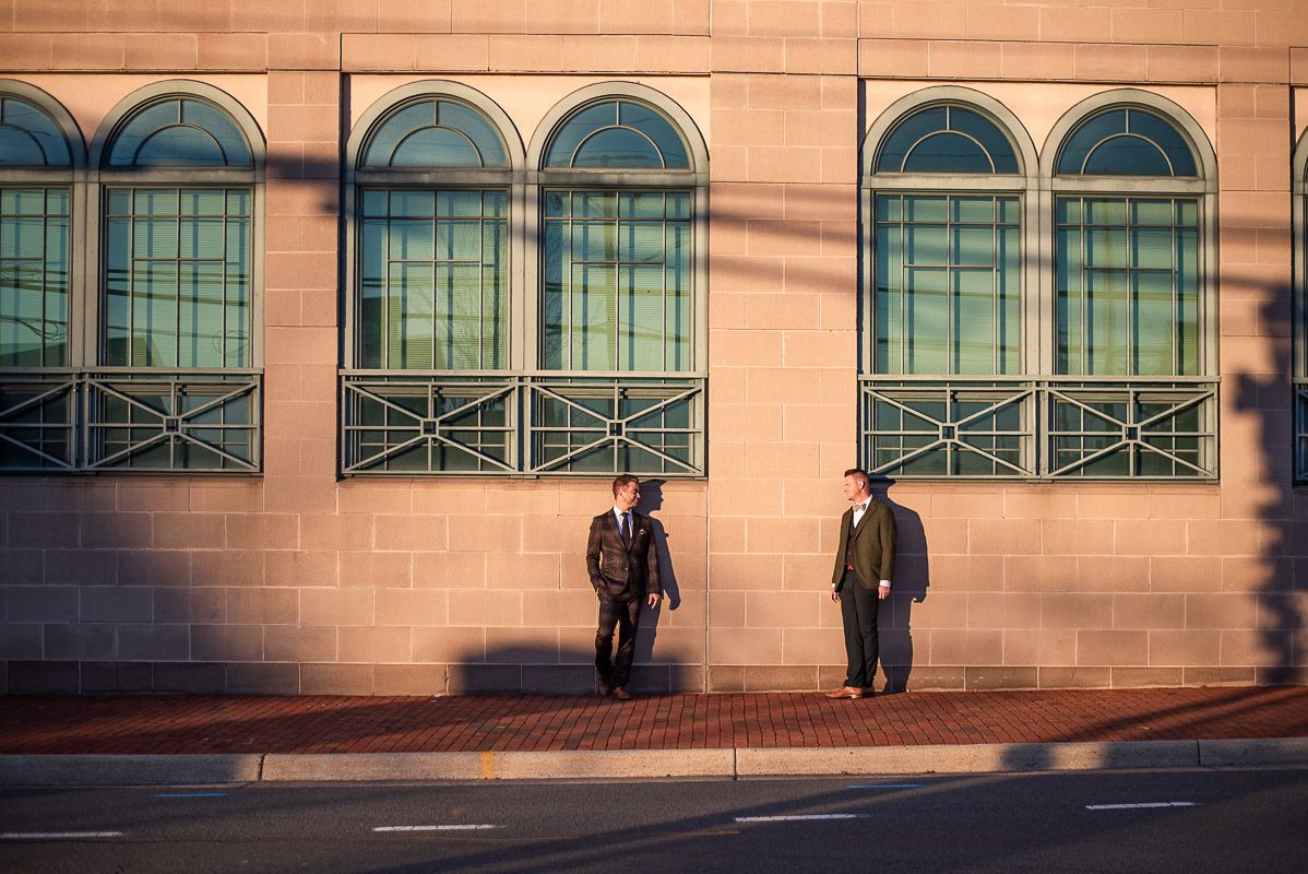 Two figures stand against a beige wall with arched green windows during sunset casting long shadows.