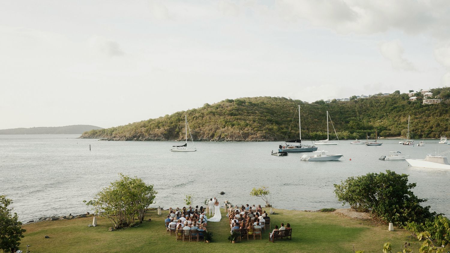 Outdoor wedding ceremony on lush green lawn overlooking a tropical bay with sailboats and verdant hillside backdrop.