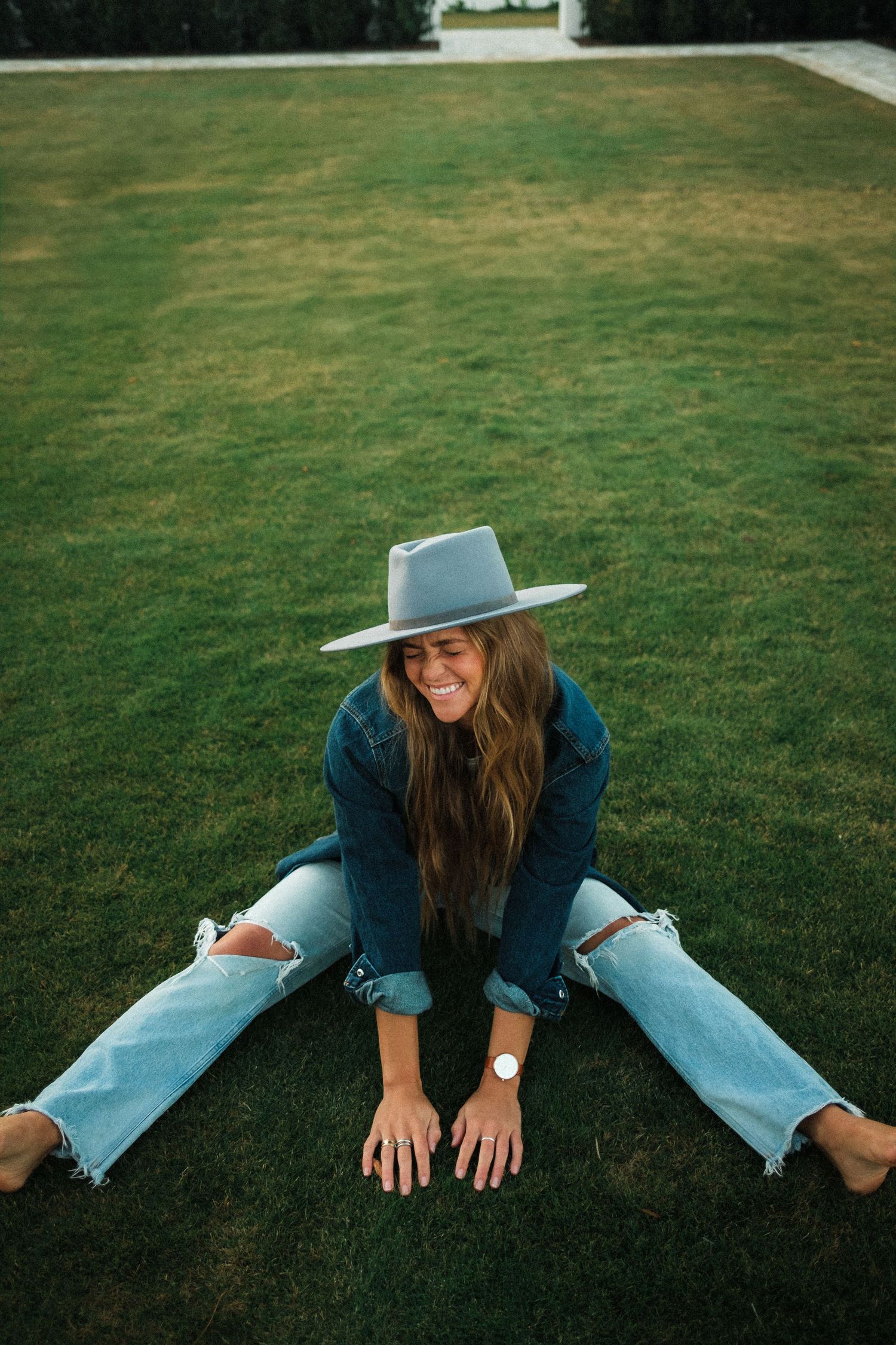A person in a blue shirt and jeans lies playfully on green grass while wearing a wide-brimmed hat.
