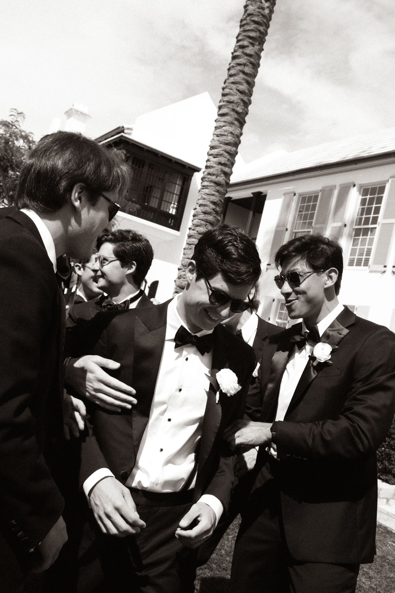 Groomsmen in black tuxedos and bow ties laughing together outdoors at a wedding celebration.