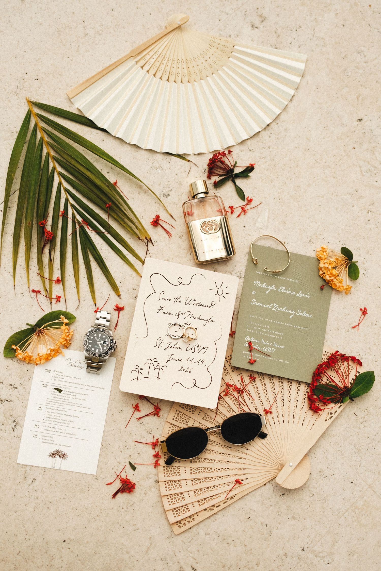 Tropical wedding flat lay with hand fans, invitations, perfume, sunglasses, watch, and floral accents on beige surface.