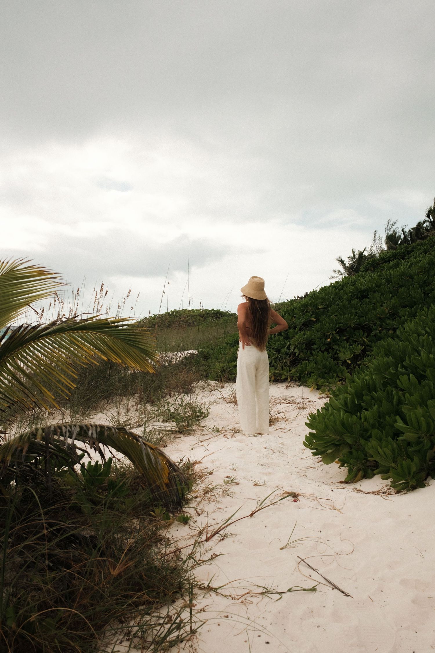 Woman in white dress and straw hat standing on sandy beach path surrounded by tropical greenery on overcast day.