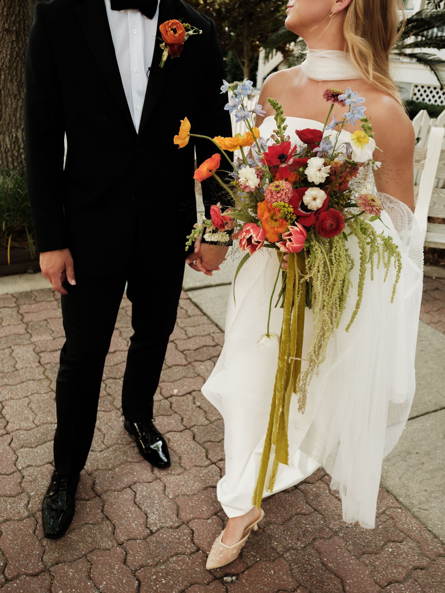 Bride holding vibrant wildflower bouquet with trailing ribbons stands beside groom in black tuxedo on brick pathway.