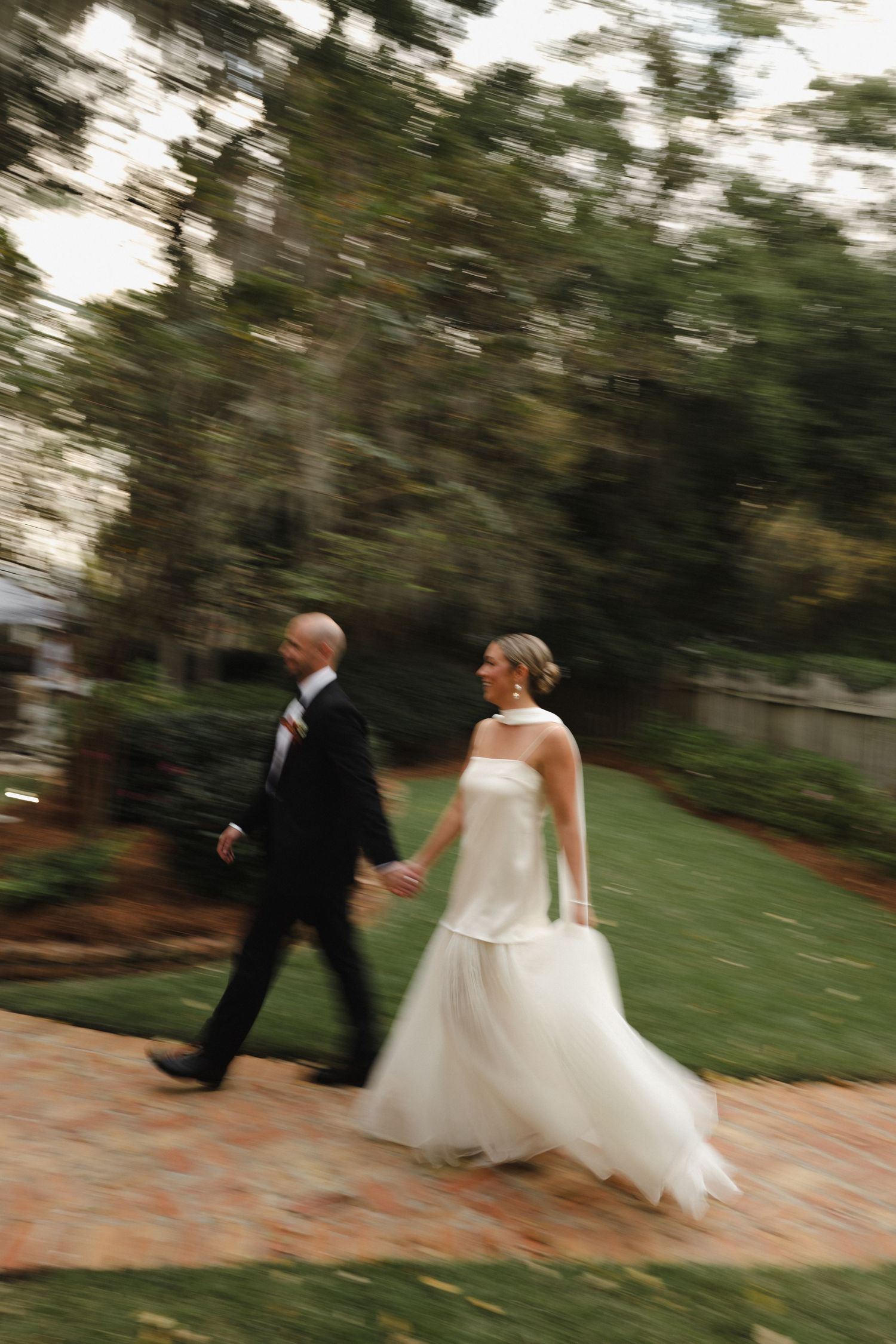 Motion blur captures a couple walking together in formal wedding attire along a garden path.