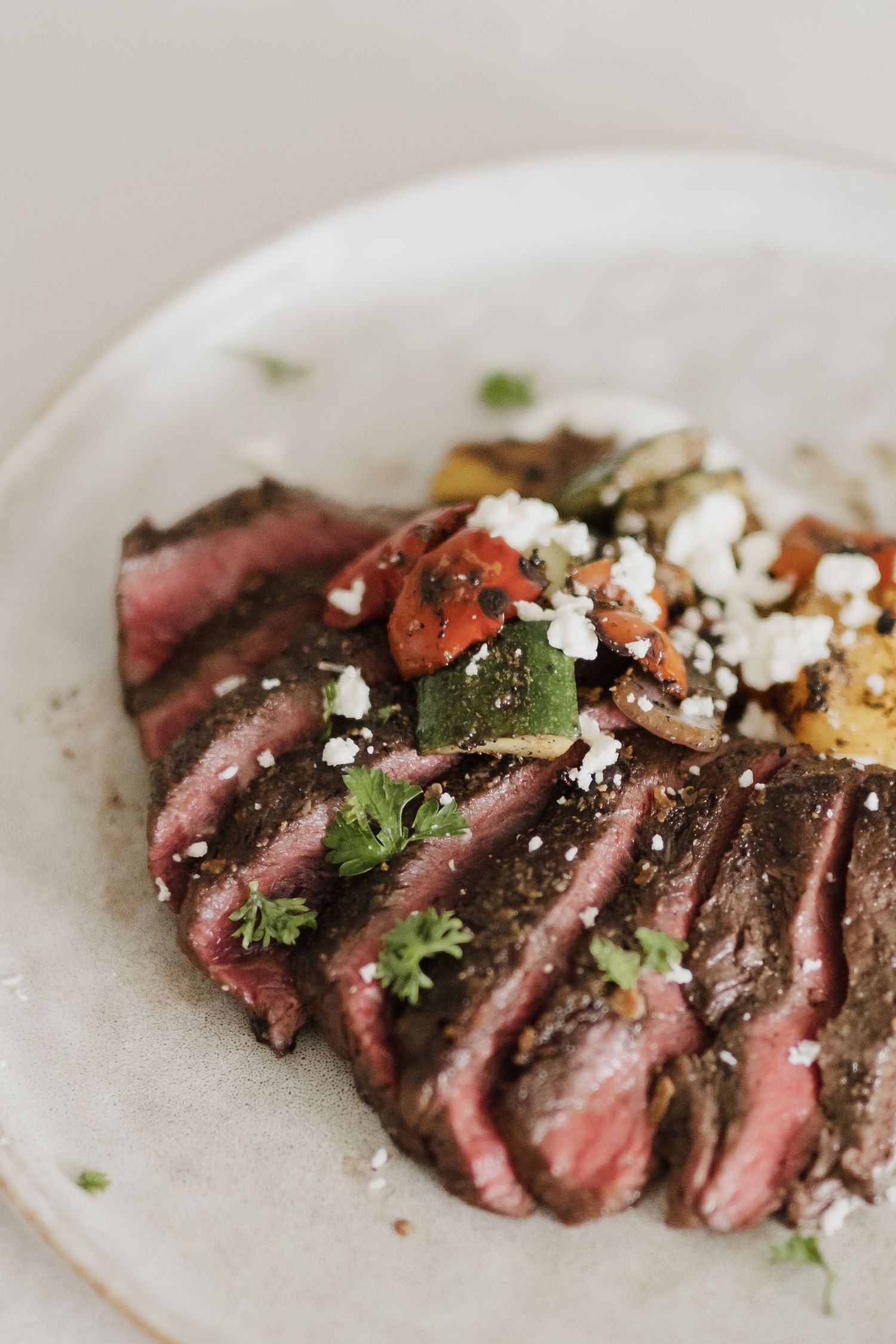 Medium-rare sliced steak topped with fresh herbs and crumbled cheese on ceramic plate.