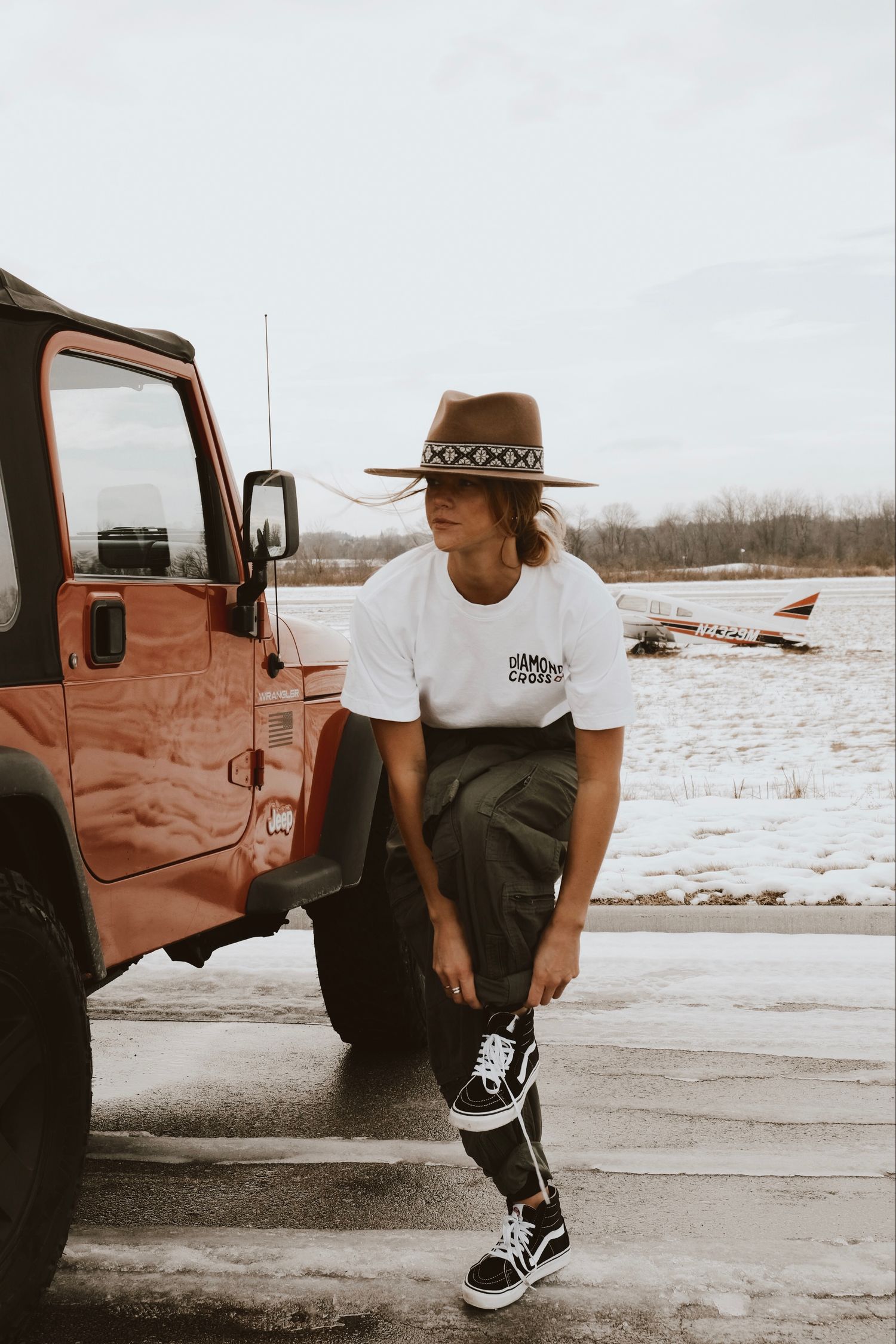 Person in brown hat, white tee, dark pants, and Vans leaning on red Jeep at snowy airfield.