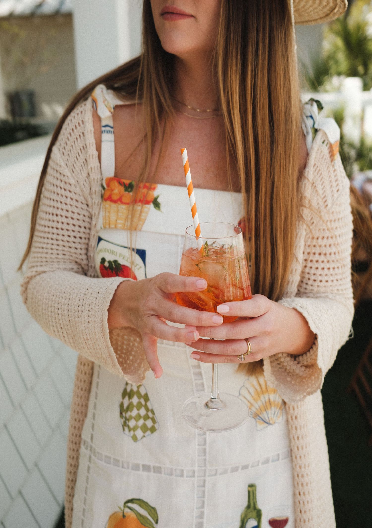 Woman in floral dress and crochet cardigan holding an Aperol spritz cocktail with orange striped straw.