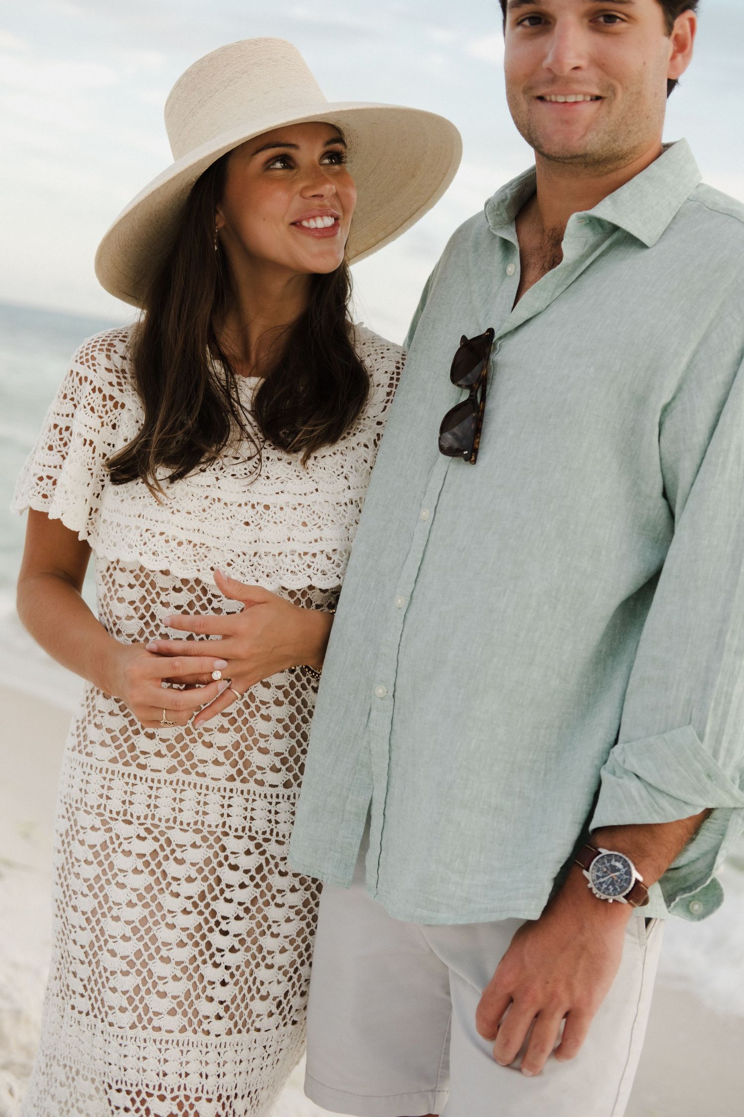 A couple poses together on a beach, wearing a white crochet dress and light blue linen shirt.