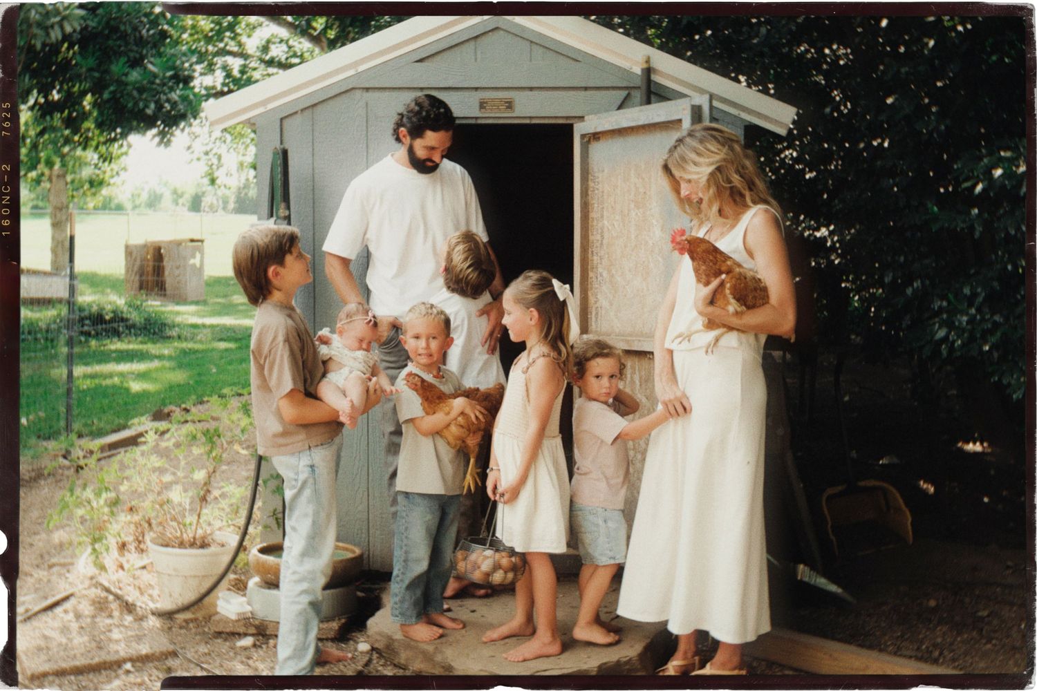 A family with several young children holds chickens outside a rustic shed on a sunny farm property.