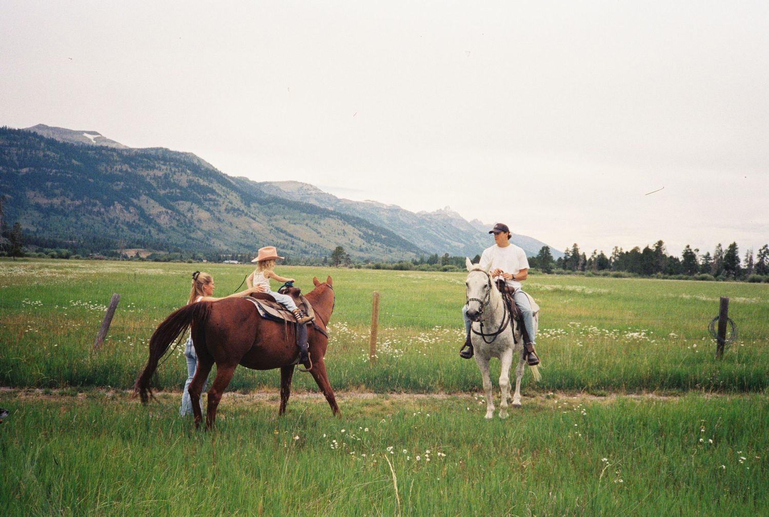 Two riders on horseback in a green meadow with mountain backdrop, wearing cowboy hats on a scenic ranch trail.