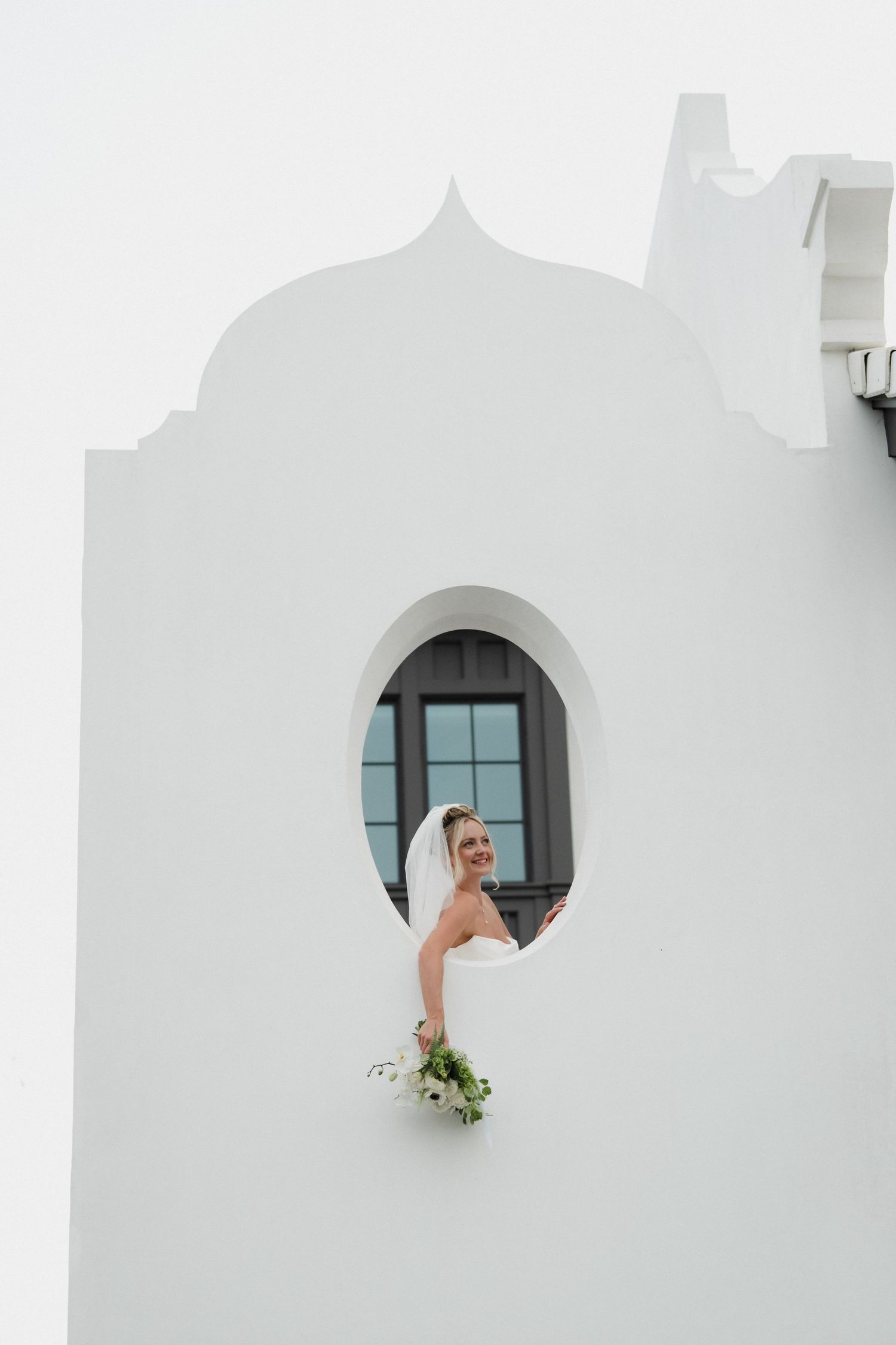 A bride in a white wedding dress poses elegantly in a circular window frame against a white stucco wall.