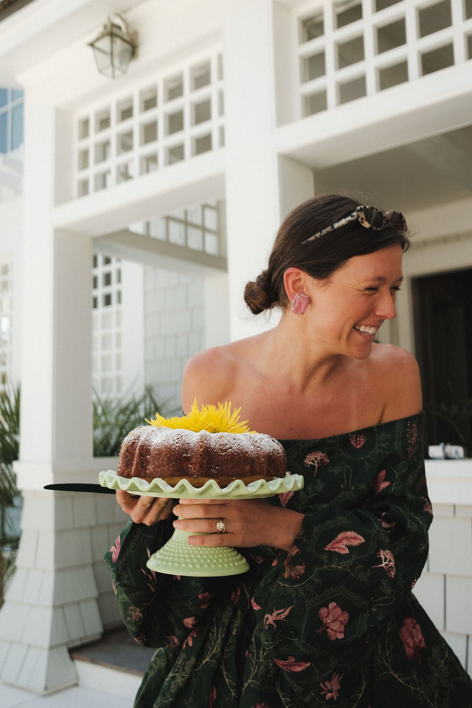 Smiling woman in floral off-shoulder dress holding a bundt cake on a green cake stand outdoors.