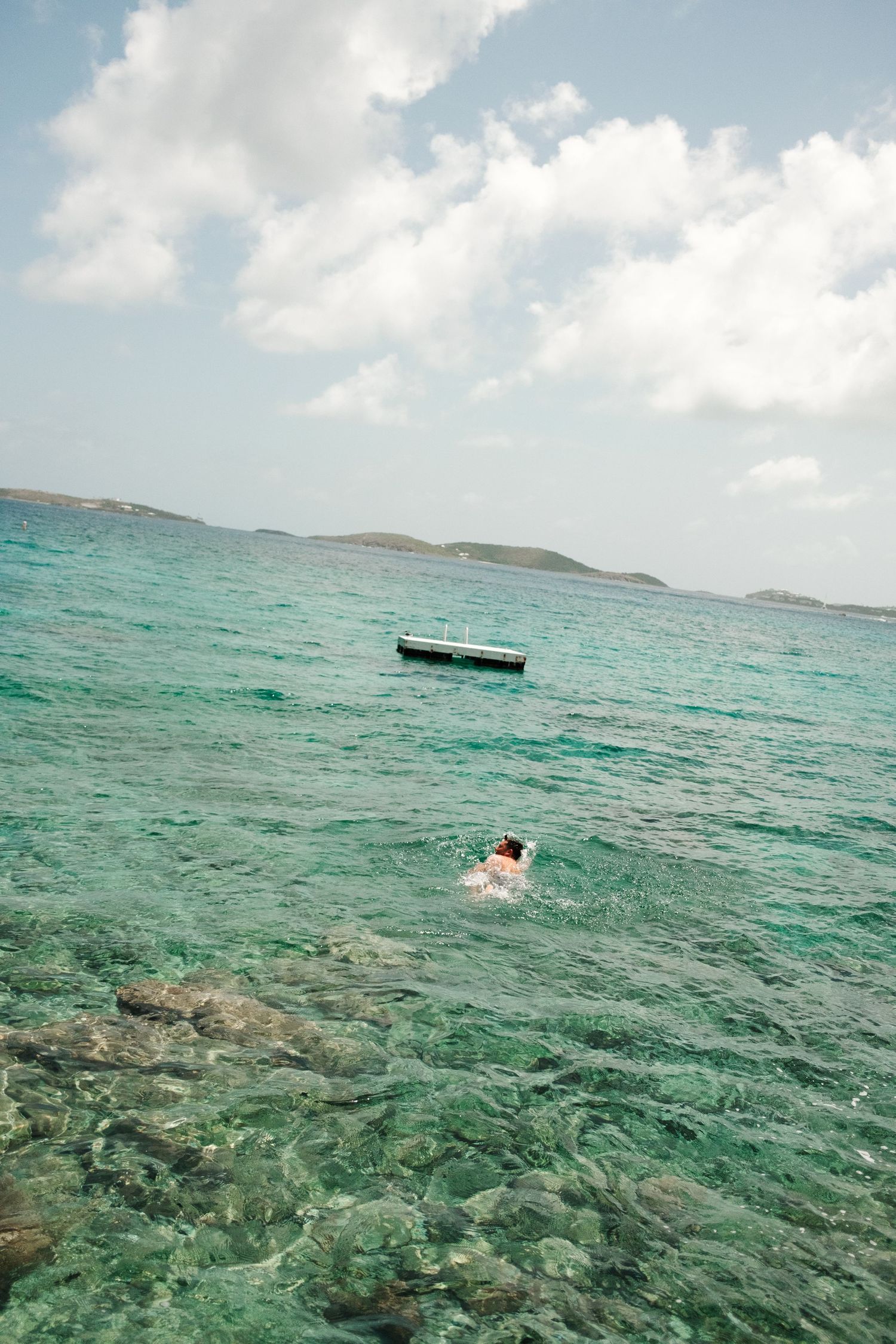 Person snorkeling in clear turquoise ocean water near rocky shore with small boat and islands in background.