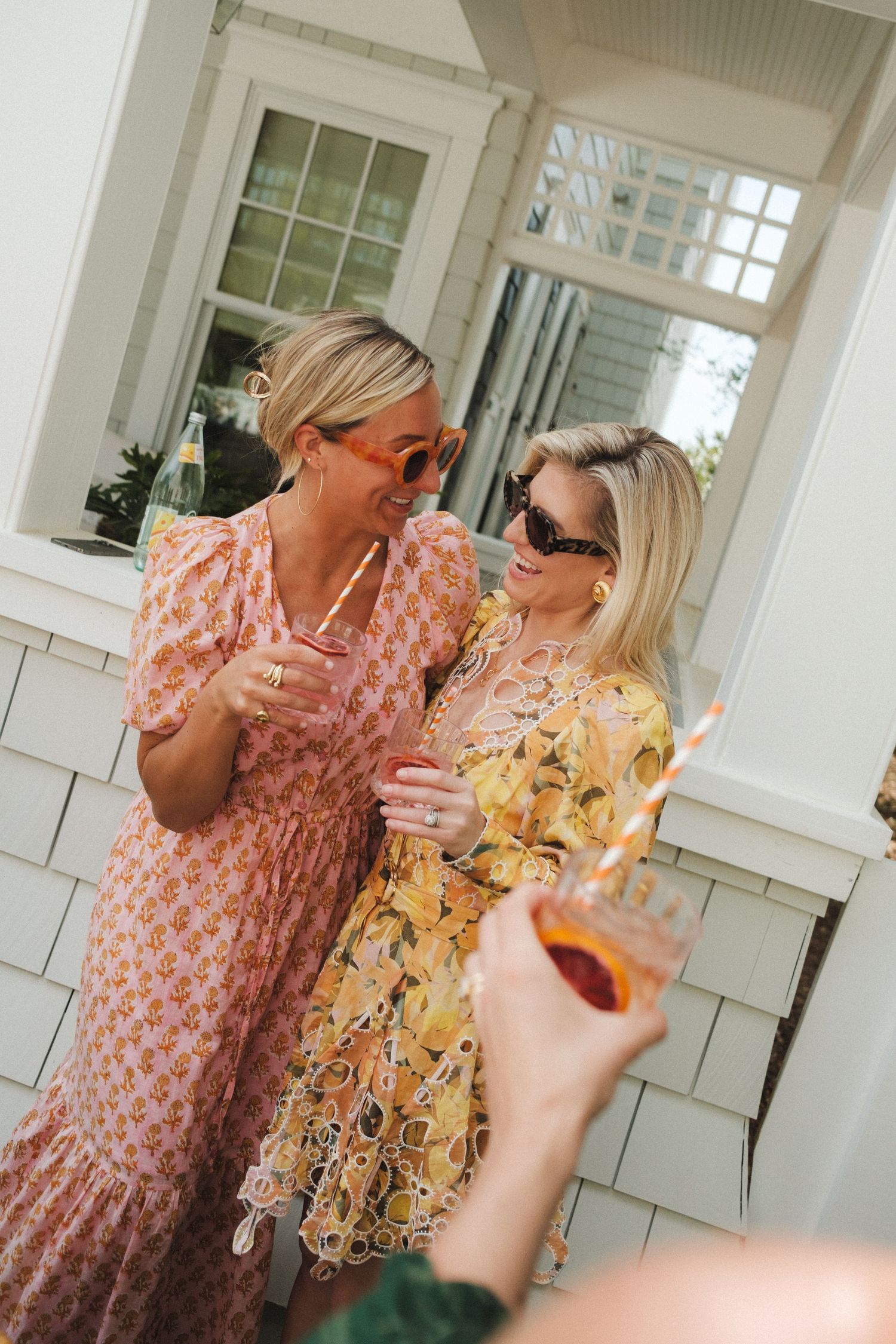 Two women in floral dresses sipping cocktails outdoors, smiling and enjoying a sunny social gathering.