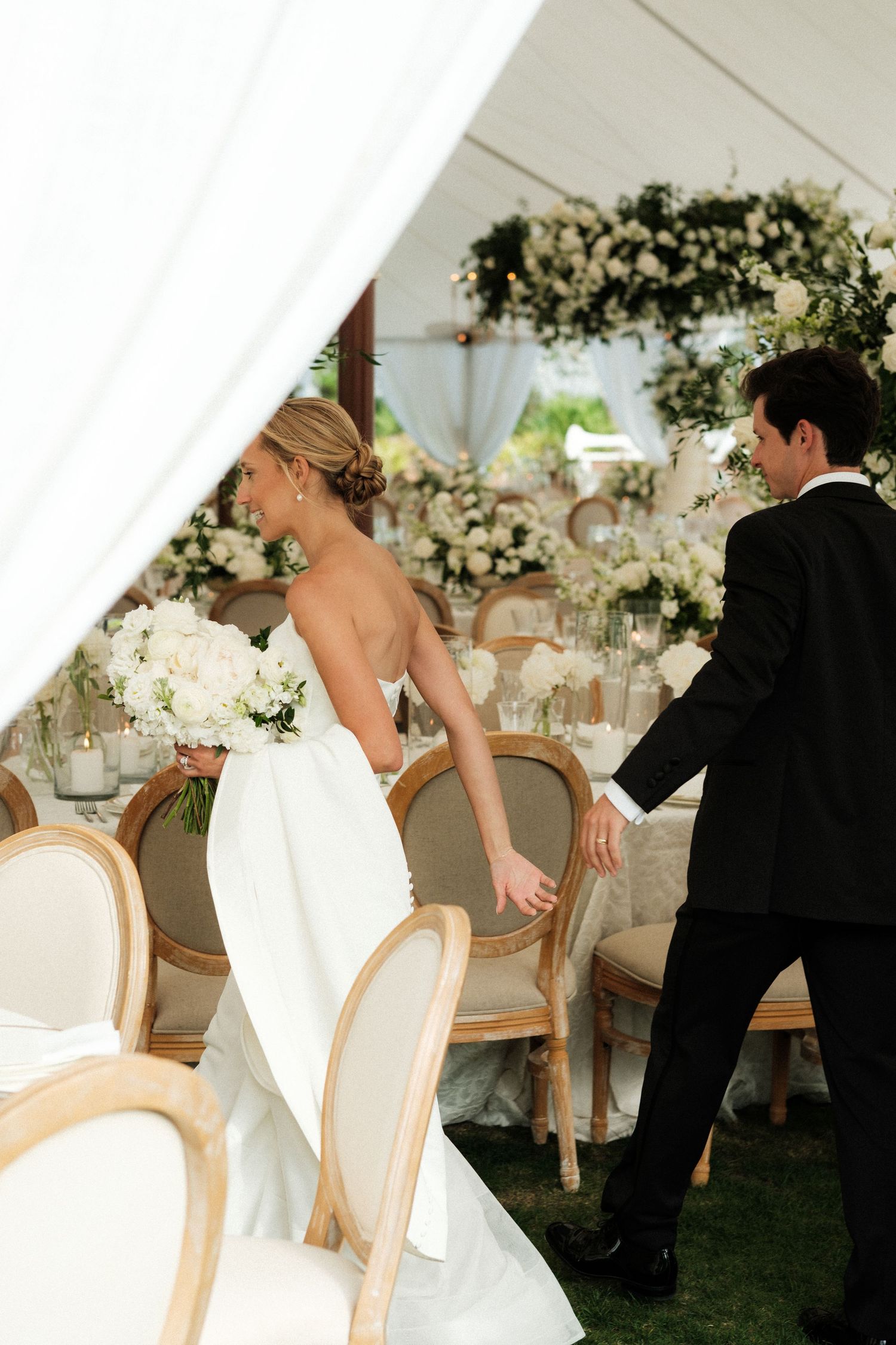Bride in white gown holding bouquet walks with groom in tuxedo into elegant floral wedding reception tent.