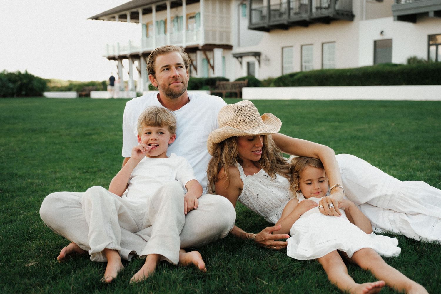 Family of four in white outfits sitting on green grass in front of a large home during a sunny day.