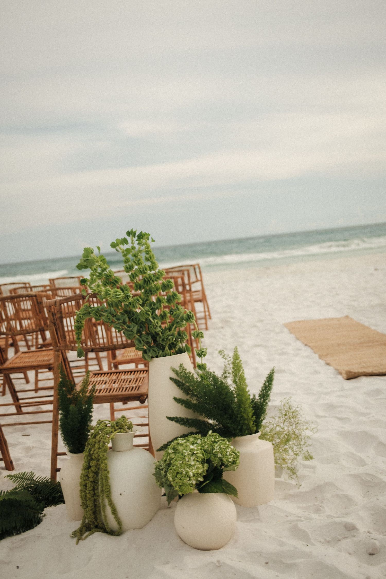 Wooden chairs and potted greenery arranged for a beach wedding ceremony with white sand and ocean waves in background.
